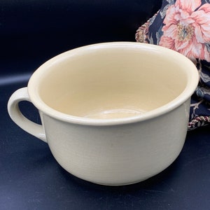 May include: A large, off-white ceramic bowl with a single handle. The bowl is round and has a smooth, slightly textured surface.