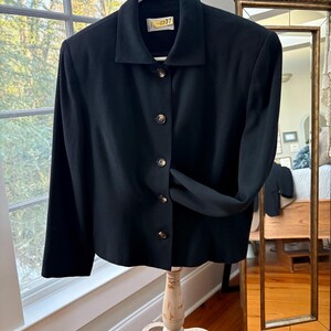 May include: A black blazer with a classic collar and gold-colored buttons. The blazer is hanging on a white hanger. The tag on the collar reads "R-77". The blazer is displayed on a vintage, distressed white stand.