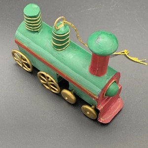 May include: A green and red wooden toy train with gold wheels and a gold string attached to the top. The train has a chimney and two stacks on top.