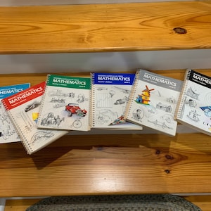 May include: A set of six spiral bound textbooks titled "Modern Curriculum Press Mathematics" with different illustrations on each cover. The books are arranged in a row on a wooden staircase.