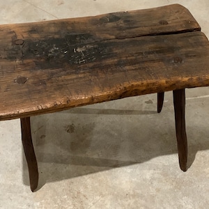 May include: A rustic wooden table with a rough-hewn top and three turned legs. The table is made of dark brown wood and has a natural, distressed finish.