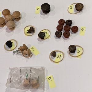 May include: Assortment of wooden and dark brown cabinet knobs, some with screws, in a variety of shapes and sizes. The knobs are arranged on a white surface with yellow sticky notes numbered 1 through 11.