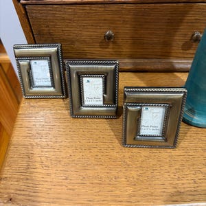 May include: Three small rectangular photo frames with a metallic bronze finish and a twisted rope border design. Each frame contains a white insert card with the text Photo Frame 1 1/2 x 1 7/8 inches printed in black ink on the center of the display.