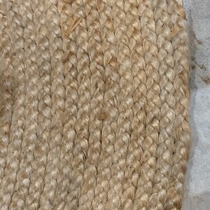 May include: Close-up of a natural fiber braided rug with a light beige color. The rug is made of tightly woven strands of natural fiber, creating a textured surface.