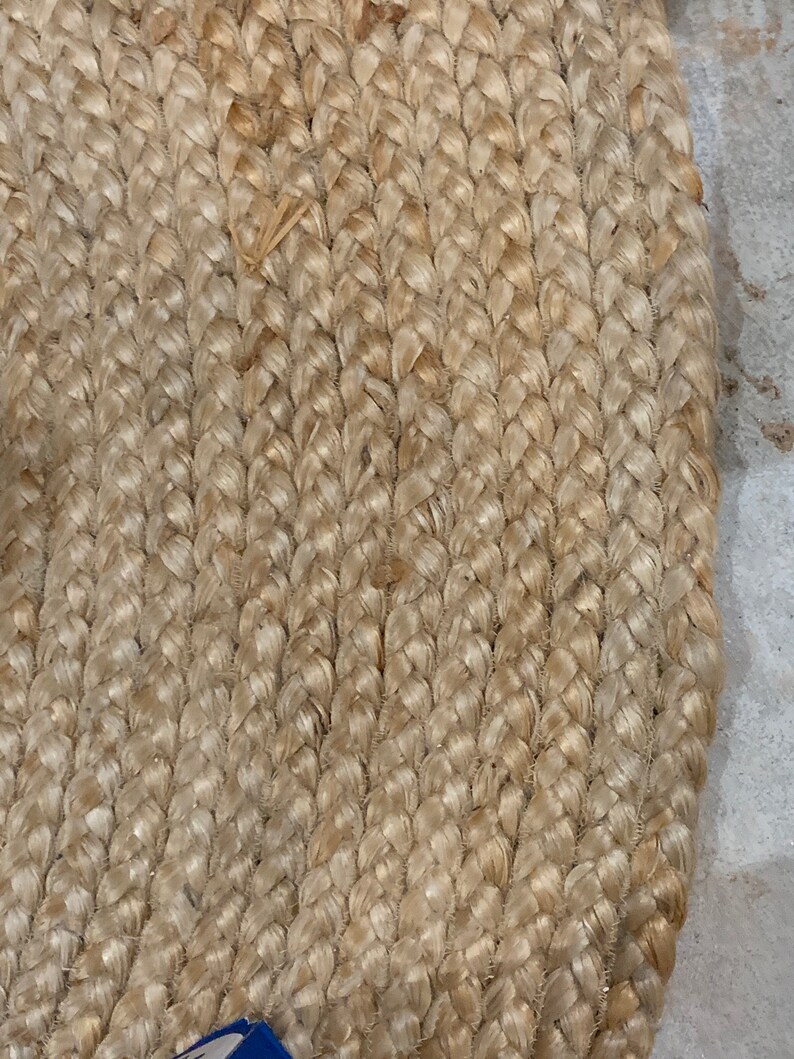 May include: Close-up of a natural fiber braided rug with a light beige color. The rug is made of tightly woven strands of natural fiber, creating a textured surface.