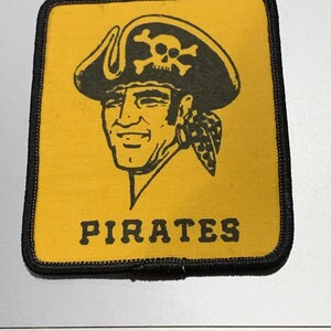 May include: A yellow fabric patch with a black border. The patch features a cartoon pirate with a skull and crossbones on his hat. The text "PIRATES" is printed in black below the pirate.