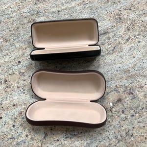 May include: Two empty eyeglass cases. One is black with a brown interior and the other is brown with a beige interior.