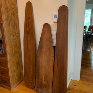 May include: Three antique wooden ironing boards of varying sizes lean against a white wall. The boards are a warm brown color, with rounded and pointed ends. The wood grain is visible, showing the natural texture of the material.