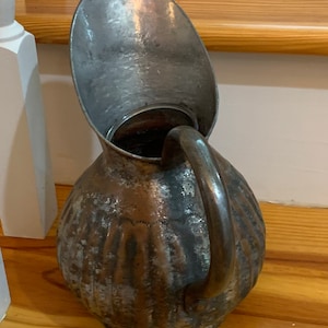 K&ouml;nnte beinhalten: Antiker Metallkrug mit gro&szlig;em, gebogenem Ausguss und Griff. Der Krug hat einen bauchigen K&ouml;rper mit strukturierter Oberfl&auml;che und einem angelaufenen, gealterten Aussehen. Der Krug ist aus Metall und wird wahrscheinlich zum Ausgie&szlig;en von Fl&uuml;ssigkeiten verwendet.