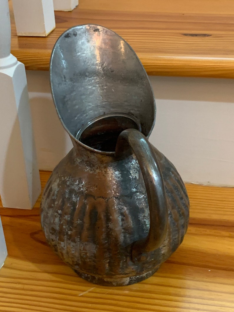 K&ouml;nnte beinhalten: Antiker Metallkrug mit gro&szlig;em, gebogenem Ausguss und Griff. Der Krug hat einen bauchigen K&ouml;rper mit strukturierter Oberfl&auml;che und einem angelaufenen, gealterten Aussehen. Der Krug ist aus Metall und wird wahrscheinlich zum Ausgie&szlig;en von Fl&uuml;ssigkeiten verwendet.