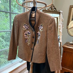 May include: A brown tweed blazer with a black collar and floral embroidery. The blazer is hanging on a wooden coat rack.