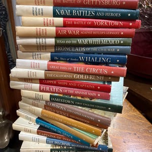 May include: A stack of vintage books from the American Heritage Junior Library series. The books have colorful covers with titles like "The Battle of Gettysburg" and "Pirates of the Spanish Main." The books are stacked on a wooden surface.