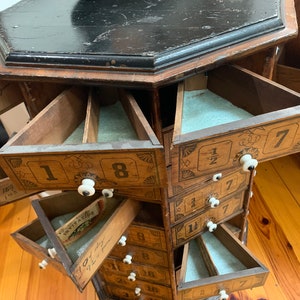 Puede incluir: Antiguo mueble de madera con m&uacute;ltiples cajones triangulares. El mueble tiene una tapa oscura, octogonal y est&aacute; pintado con n&uacute;meros y elementos decorativos. Cada caj&oacute;n tiene un pomo blanco y est&aacute; parcialmente abierto.