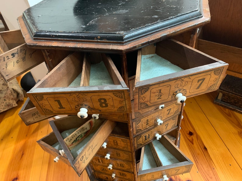Puede incluir: Antiguo mueble de madera con m&uacute;ltiples cajones triangulares. El mueble tiene una tapa oscura, octogonal y est&aacute; pintado con n&uacute;meros y elementos decorativos. Cada caj&oacute;n tiene un pomo blanco y est&aacute; parcialmente abierto.