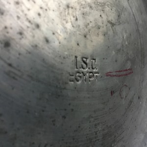 K&ouml;nnte beinhalten: Nahaufnahme einer metallischen Oberfl&auml;che mit dem eingestempelten Text "I.S.C. EGYPT". Das Metall hat ein abgenutztes, strukturiertes Aussehen mit sichtbaren Kratzern und Unvollkommenheiten. Eine rote Markierung ist vorhanden.
