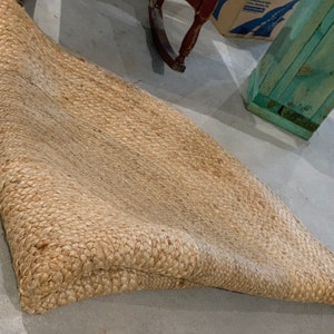 May include: A natural fiber rug made of woven jute, with a light brown color and a slightly textured surface. The rug is folded in half and lying on a concrete floor.