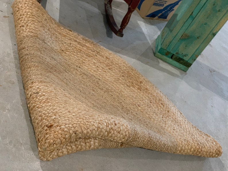 May include: A natural fiber rug made of woven jute, with a light brown color and a slightly textured surface. The rug is folded in half and lying on a concrete floor.