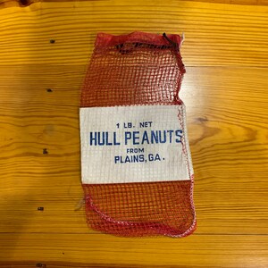 May include: A red mesh bag with a white label that reads "1 LB. NET HULL PEANUTS FROM PLAINS, GA."