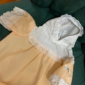 May include: A pale peach baby gown with white lace trim and a white lace collar. The gown has a small label that says "First Impressions".