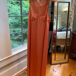 May include: A long, peach-colored slip dress with a lace trim. The dress is hanging on a wooden hanger.
