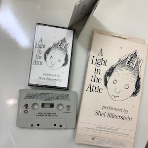 May include: A cassette tape and a cardboard box with a black and white illustration of a person with a house on their head. The text on the cassette tape reads "Shel Silverstein A Light in the Attic" and the text on the box reads "A Light in the Attic performed by Shel Silverstein Special Package Contains One Cassette & Selected Poems & Drawings".
