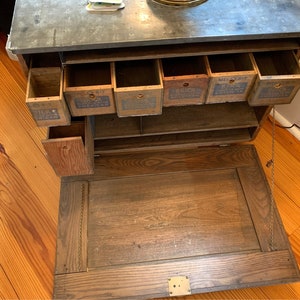 May include: Antique wooden cabinet with a gray countertop and multiple drawers. The drawers have labels that read "Milk Bit", "Brick", "Mello-Rit", and "Brick". The cabinet has a drop-down door.
