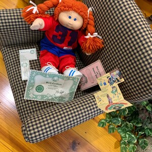 May include: A Cabbage Patch Kids doll with red yarn pigtails, wearing a red and blue football uniform with the number 31. The doll is seated on a checkered armchair, with birth certificates and other ephemera.