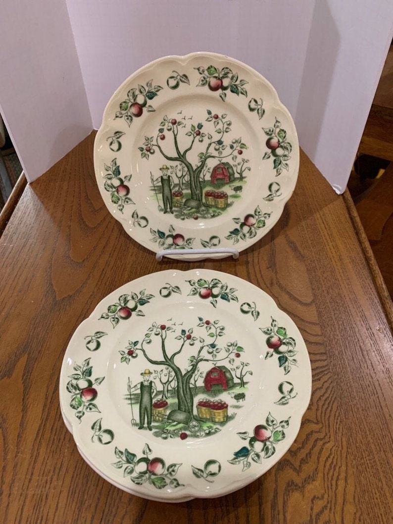 May include: Set of three white ceramic dinner plates with a green, red, and brown apple orchard design. The plates feature a stylized apple tree with red apples, a red barn, and a person picking apples.