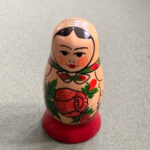 May include: A traditional wooden nesting doll, also known as a matryoshka doll, painted with a floral design. The doll is wearing a green and red scarf and has a red and white floral pattern on its dress. The doll has a red base.