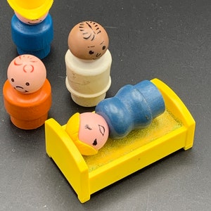 May include: A set of four vintage plastic Little People figures, including a yellow-hatted figure, a brown-skinned figure, a figure with a red and white face, and a figure with blonde hair lying in a yellow bed.