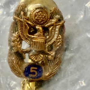 May include: Gold-toned pin with an eagle and a blue circle with the number 5. The eagle is holding a shield with a star in the center. The pin has a chain attached to the back.
