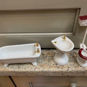 May include: A set of three miniature white porcelain bathroom fixtures, including a bathtub, a sink, and a toilet. The bathtub and sink have gold-colored faucets. The toilet has a red lid and a gold chain.