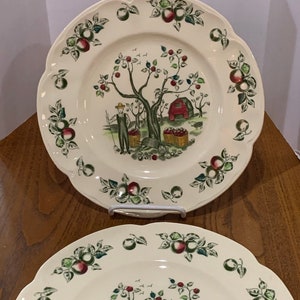 May include: Two white ceramic dinner plates with a scalloped edge. The plates feature a green, red, and brown apple tree design with a red barn and a person picking apples. The border of the plates is decorated with green leaves and red apples.
