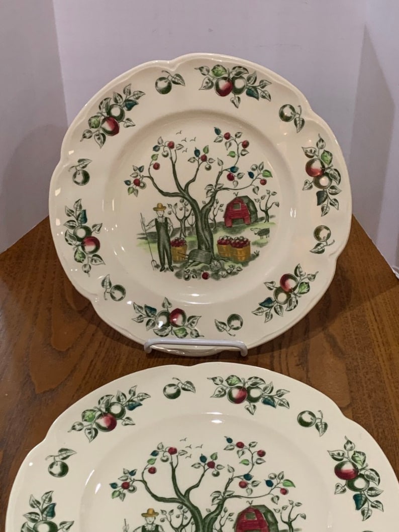 May include: Two white ceramic dinner plates with a scalloped edge. The plates feature a green, red, and brown apple tree design with a red barn and a person picking apples. The border of the plates is decorated with green leaves and red apples.