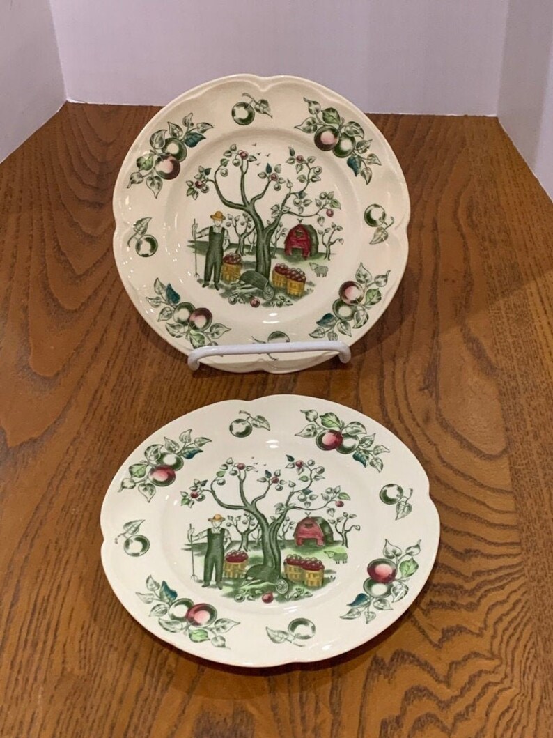 May include: Two white ceramic plates with a green, red, and brown apple orchard design. The plates feature a man picking apples from a tree and a small red barn.