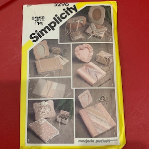 May include: Simplicity sewing pattern number 5296 for making six different types of small boxes. The boxes are decorated with lace, ribbon, and other embellishments. The pattern is designed by Marjorie Puckett.