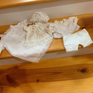 May include: A set of white lace doll clothes, including a dress, a top, a pair of pants, and a bonnet. The clothes are laid out on a wooden staircase.