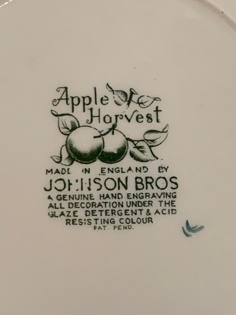 May include: A white ceramic plate with a green and black design. The design features a stylized apple with leaves and the text "Apple for Harvest" and "Johnson Bros". The text also includes "Made in England" and "A genuine hand engraving all decoration under the glaze detergent & acid resisting colour Pat. Pend."