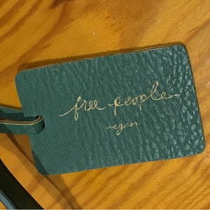 Puede incluir: Una etiqueta de cuero verde oscuro con las palabras "free people" en escritura dorada y la palabra "vegan" debajo. La etiqueta est&aacute; sujeta a una correa de cuero verde oscuro. El fondo es una superficie de madera.