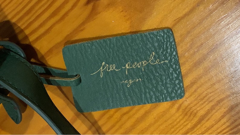 Puede incluir: Una etiqueta de cuero verde oscuro con las palabras "free people" en escritura dorada y la palabra "vegan" debajo. La etiqueta est&aacute; sujeta a una correa de cuero verde oscuro. El fondo es una superficie de madera.