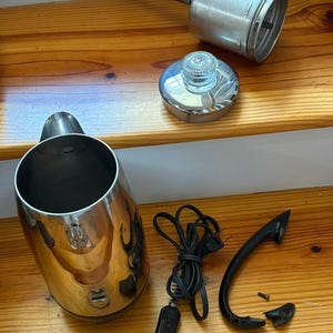 May include: A disassembled vintage chrome electric kettle with a detached lid, handle, and power cord. The kettle's components are arranged on a wooden surface, showcasing its parts.