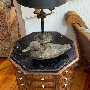 Puede incluir: Un mueble octogonal antiguo con cajones, coronado con una superficie negra, una l&aacute;mpara con una pantalla negra y un se&ntilde;uelo de pato de madera. El mueble tiene pomos blancos y cajones numerados.