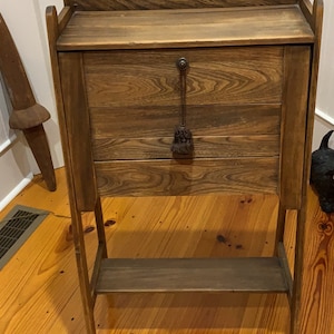 May include: A dark brown wooden secretary desk with a drop-down front and a lower shelf. The desk has a decorative metal pull with a tassel.