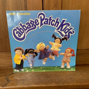 May include: 1985 Cabbage Patch Kids calendar featuring five dolls jumping rope against a blue sky background. The dolls are wearing colorful clothing and have different hair colors and styles.