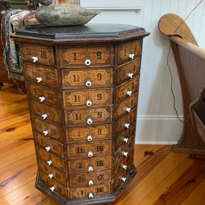 Puede incluir: Un antiguo mueble de farmacia de madera con m&uacute;ltiples cajones, cada uno etiquetado con n&uacute;meros y fracciones. El mueble tiene un acabado oscuro, pomos blancos y una tapa negra. Un se&ntilde;uelo de pato de madera descansa encima. El mueble es de forma octogonal.