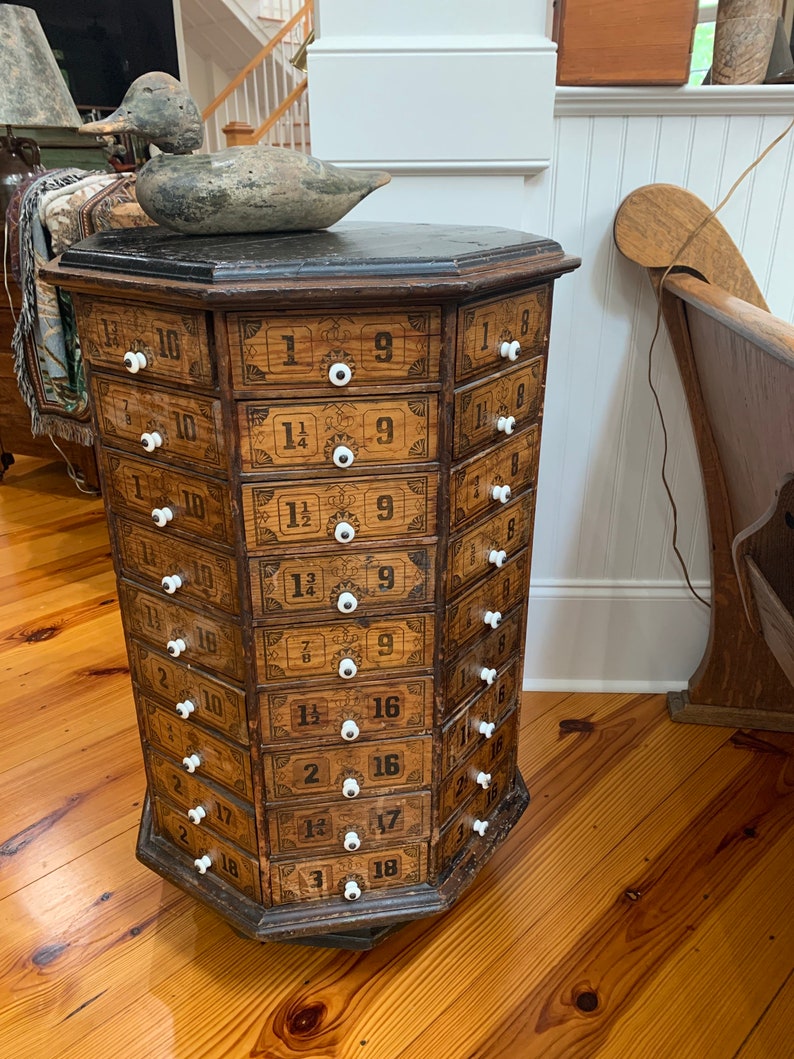 Puede incluir: Un antiguo mueble de farmacia de madera con m&uacute;ltiples cajones, cada uno etiquetado con n&uacute;meros y fracciones. El mueble tiene un acabado oscuro, pomos blancos y una tapa negra. Un se&ntilde;uelo de pato de madera descansa encima. El mueble es de forma octogonal.