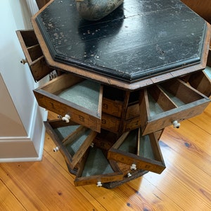 Puede incluir: Un mueble octogonal antiguo de madera con m&uacute;ltiples cajones, cada uno numerado. El mueble tiene una tapa negra y un aspecto envejecido. Un se&ntilde;uelo de pato de madera se encuentra encima. Los cajones tienen pomos blancos y est&aacute;n parcialmente abiertos.