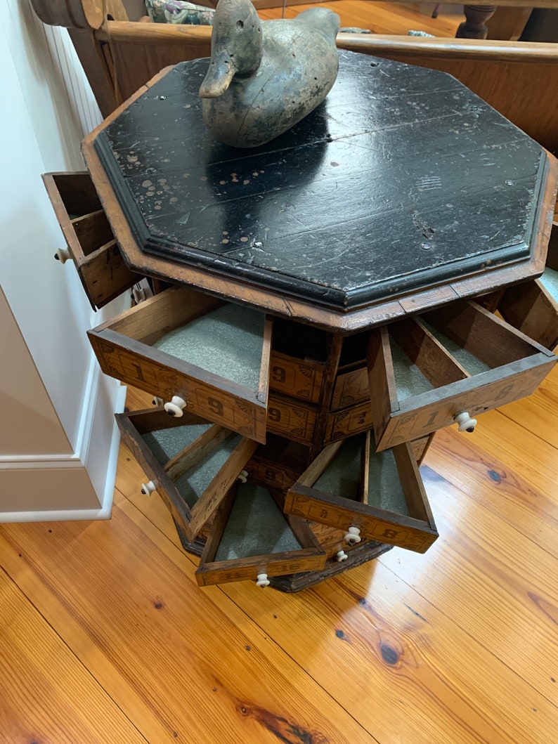 Puede incluir: Un mueble octogonal antiguo de madera con m&uacute;ltiples cajones, cada uno numerado. El mueble tiene una tapa negra y un aspecto envejecido. Un se&ntilde;uelo de pato de madera se encuentra encima. Los cajones tienen pomos blancos y est&aacute;n parcialmente abiertos.