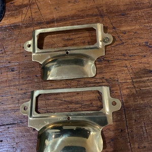 May include: Two brass drawer pulls with a rectangular label holder. The pulls have a rounded, curved shape and are designed to be used on drawers or cabinets.
