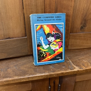 May include: A blue book with the title "The Campfire Girls Flying Around the Globe" and a colorful illustration of a girl flying a plane.
